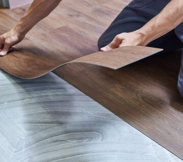 A person installs vinyl flooring, carefully aligning a dark wood-patterned vinyl plank over an adhesive spread on a concrete subfloor, ensuring it seamlessly complements the durability of epoxy floor coatings.