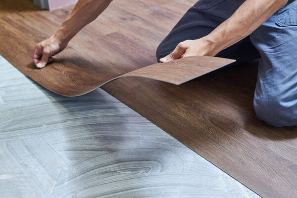 A person installs vinyl flooring, carefully aligning a dark wood-patterned vinyl plank over an adhesive spread on a concrete subfloor, ensuring it seamlessly complements the durability of epoxy floor coatings. A person installs vinyl flooring, carefully aligning a dark wood-patterned vinyl plank over an adhesive spread on a concrete subfloor, ensuring it seamlessly complements the durability of epoxy floor coatings.