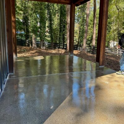 Polished concrete floor in a wooden outdoor area with trees and a fence in the background. A person stands partially visible on the right. Polished concrete floor in a wooden outdoor area with trees and a fence in the background. A person stands partially visible on the right.